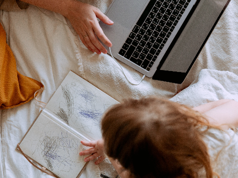 a toddler scribbling in a notebook near an open laptop
